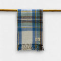 A plaid patterned scarf in stewart muted blue, green, and beige, displayed on a horizontal rod, with a label showing the brand name.