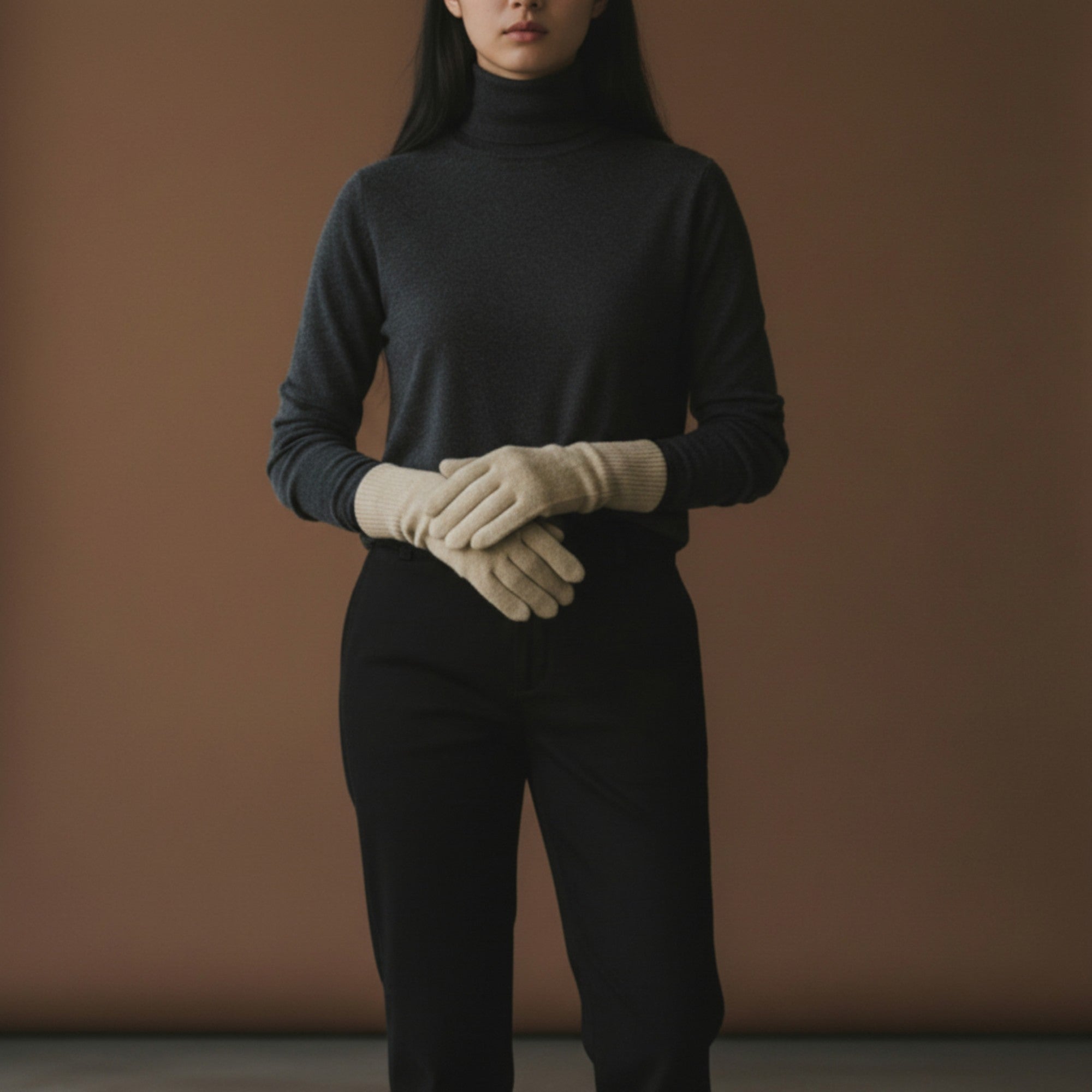 Person wearing a dark turtleneck and black pants with beige gloves against a brown background